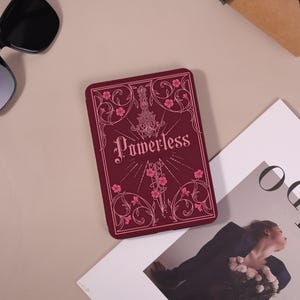 May include: A rectangular e-reader case in deep burgundy featuring a central graphic of a sword surrounded by intricate floral line art and the word Powerless printed in a stylized font. The design includes delicate pink flower accents.