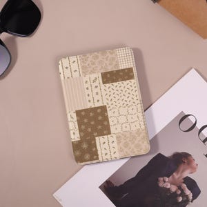 Może przedstawiać: Prostokątne etui z patchworkowym wzorem w odcieniach beżu, brązu i kremu. Wzór zawiera elementy kwiatowe i geometryczne. Etui leży na jasnobrązowej powierzchni, z okularami przeciwsłonecznymi i magazynem w tle.