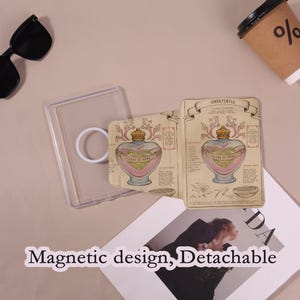 May include: A clear phone case with a white ring grip and a decorative card featuring a heart-shaped potion bottle design with the text "FILTRE DE AMOR". The card has a vintage aesthetic with the word "AMORTENTIA" at the top. The text "Magnetic design, Detachable" is also visible.