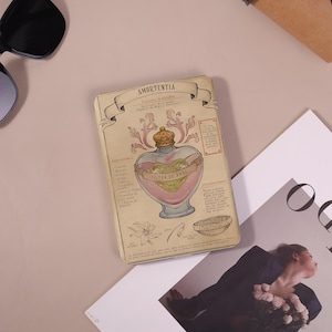 May include: A vintage-style notebook with the word "AMORTENTIA" at the top. The cover features a pink and gold heart-shaped bottle with the words "FILTRO DE AMOR" and botanical illustrations. The notebook has a cream-colored background.