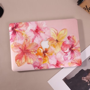 May include: A pink laptop case adorned with a watercolor floral design. The case features a pattern of hibiscus flowers in shades of pink, orange, and yellow. The Apple logo is visible in the center. The case is designed to fit a laptop.