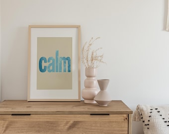May include: A framed print with the word "calm" in blue watercolor lettering. The print is displayed on a wooden dresser, alongside two decorative vases and dried plants. The scene evokes a sense of tranquility.