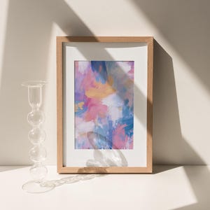 May include: A framed abstract art print with a wooden frame. The artwork features a colorful blend of pink, blue, and gold brushstrokes. A clear glass candle holder with a round base and a series of spherical accents is to the left.