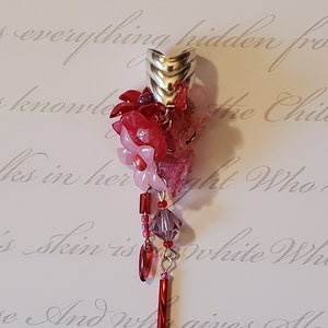 Può includere: Un ciondolo fatto a mano con un grappolo di fiori rossi e rosa, impreziosito da perline rosse e viola e una lunga perla rettangolare rossa. Il ciondolo ha una parte superiore in metallo color argento.