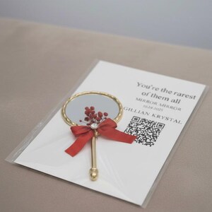Pu&ograve; includere: Un piccolo specchio a mano rotondo dorato con un fiocco rosso e decorazioni floreali. Lo specchio &egrave; su un biglietto bianco con la scritta "You're the rarest of them all".