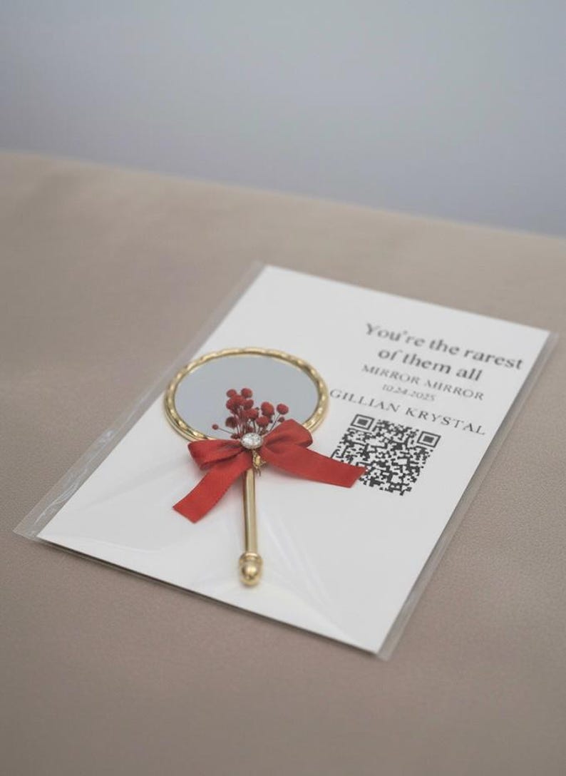 Pu&ograve; includere: Un piccolo specchio a mano rotondo dorato con un fiocco rosso e decorazioni floreali. Lo specchio &egrave; su un biglietto bianco con la scritta "You're the rarest of them all".