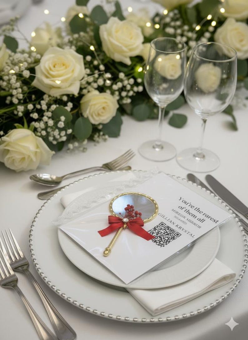 Pu&ograve; includere: Un'apparecchiatura per la tavola nuziale con rose bianche, verde e due calici. Un piccolo specchio a mano con un fiocco rosso e accenti floreali poggia su un piatto bianco con posate e un tovagliolo. La carta recita "You're the rarest of them all".