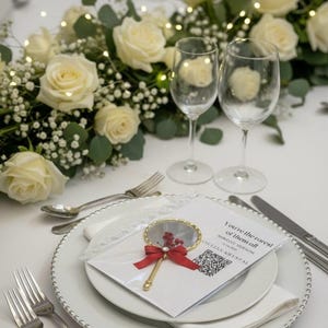 Pu&ograve; includere: Un'impostazione del tavolo nuziale con rose bianche, verde e luci fatate. Due calici sono vicino a un piatto con un segnaposto decorativo e una carta con la scritta "You're the rarest of them all". Posate d'argento e una tovaglia bianca.