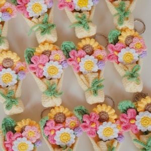 Pode incluir: Uma coleção de buquês de flores de crochê feitos à mão em várias cores, incluindo girassóis amarelos, margaridas rosa e brancas e detalhes roxos. Cada buquê é embrulhado em um cone amarelo claro e tem folhas verdes.