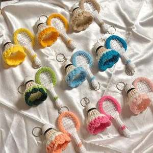 May include: A collection of colorful, handmade badminton shuttlecock keychains. Each keychain features a crocheted shuttlecock with a matching racket, available in various colors like yellow, orange, green, blue, and pink. The keychains are attached to silver keyrings.