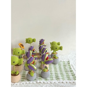 May include: A collection of handmade crochet plants in small pots. The plants feature green, purple, and yellow colors, with some resembling carnivorous plants. The pots are in various neutral colors, and the background is a white wall and a green and white checkered tablecloth.