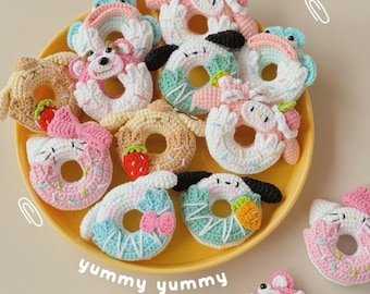 May include: A selection of handmade crocheted donuts in various colours and designs, featuring animal and fruit motifs. The donuts, in shades of white, pink, blue, and brown, are displayed on a yellow plate, with the words "yummy yummy" visible.