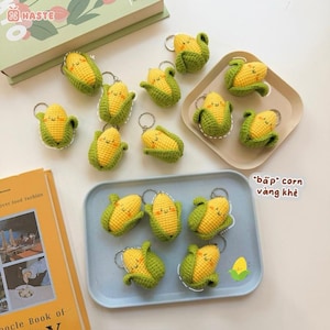 May include: A selection of crocheted corn keyrings. Each keyring features a yellow corn cob with green husks and a smiling face. The keyrings are arranged on a light blue tray and a beige tray, with a book and a box in the background.