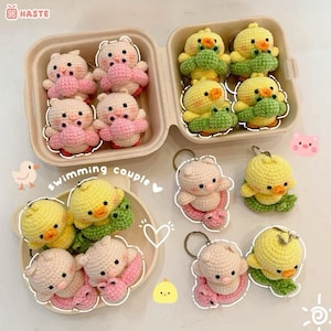 以下が含まれることがあります： 愛らしい動物のデザインが特徴の手作りのかぎ針編みキーホルダーのコレクション。キーホルダーには、ピンクの豚と緑のアクセントが付いた黄色いアヒルが含まれており、容器とプレートに配置されています。「swimming couple」というテキストも表示されます。