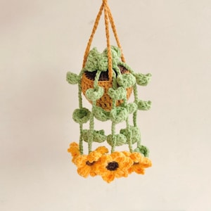 May include: A handmade crochet hanging planter with a woven basket design. The planter is filled with green crochet leaves and adorned with a ring of orange and brown crochet sunflowers. The hanging cord is a golden brown color.