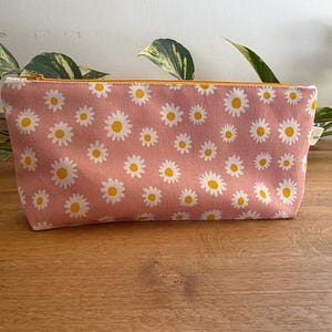 May include: A pink zippered pouch with a white daisy pattern. The pouch has a yellow zipper.