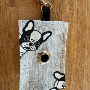 Könnte beinhalten: Grauer Stoff-Kotbeutelspender mit einem schwarz-weißen Französisch-Bulldoggen-Design. Der Spender hat eine Metallöse und einen silberfarbenen Clip zum Befestigen an einer Leine. Eine schwarze Stoffschlaufe ist am Clip befestigt.