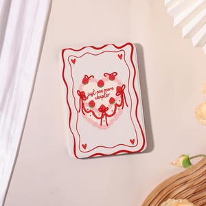 May include: A white rectangular case with a red border and heart accents. The case features a heart-shaped design with strawberries, bows, and the text "just one more chapter".