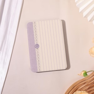 May include: A rectangular notebook with a light purple fabric spine and a cream-colored cover featuring thin vertical blue lines. A strip of white lace trim runs vertically along the spine, accented by a single small, round, light purple button.
