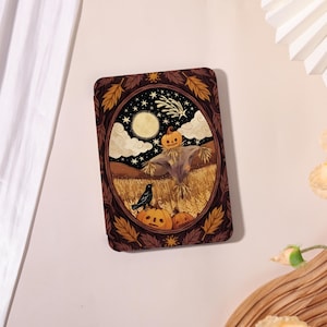 May include: A rectangular e-reader case featuring an oval illustration of a scarecrow with a pumpkin head in a wheat field under a night sky with a full moon, stars, and a shooting star, framed by a border of brown and gold autumn leaves.