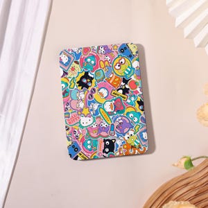 Könnte beinhalten: Ein rechteckiges, farbenfrohes Etui, das mit Cartoon-Aufklebern bedeckt ist. Die Aufkleber zeigen verschiedene Charaktere in leuchtenden Farben wie Pink, Blau, Gelb und Grün. Das Etui hat eine glatte Oberfläche und eine abgerundete Kante.