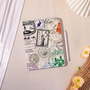 May include: A white tablet case with a design of vintage travel stamps from places like New York, Rome, and Mexico. A white stylus is placed on the side. The case has a cream-colored background.