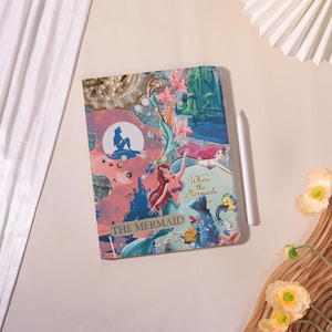 Może przedstawiać: Dekoracyjny notatnik z kolażem ilustracji z "Małej Syrenki". Okładka przedstawia Ariel, Florka i Sebastiana, z tekstem "Where the Mermaids are" i "The Mermaid". Obok leży biały rysik.