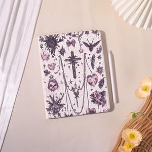 May include: A white tablet case with a gothic design featuring crosses, hearts, skulls, and chains in black and pink. A white stylus rests beside the case. The case has a rectangular shape.