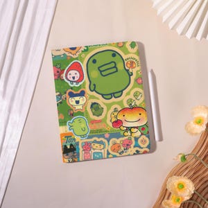 Peut inclure: Une coque de tablette colorée avec des personnages de dessins animés et le texte "Kuchipatchi". L'étui a un fond vert avec divers autocollants de personnages et un stylet blanc.