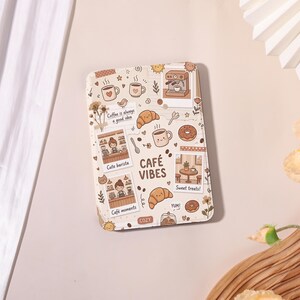 May include: A rectangular tablet case featuring a cream-colored background with brown illustrations of coffee, croissants, donuts, and a barista. The text reads "Coffee is always a good idea," "Cute barista," "Café moments," "CAFÉ VIBES," "Sweet treats," "YUM!," and "COZY."