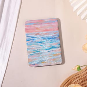 May include: A rectangular art piece with rounded corners, featuring an impressionistic seascape. The artwork depicts a sunset over the ocean, with hues of pink, blue, and white. The brushstrokes create a textured effect, evoking a sense of calm.