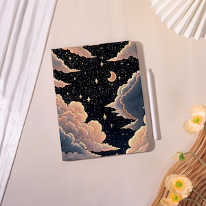 May include: A notebook with a night sky design featuring clouds, stars, and a crescent moon. The cover has a dark blue background with peach and blue clouds. A white stylus rests beside the notebook.