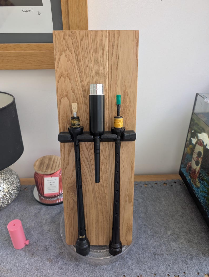 May include: Three black bagpipes with a black and silver cleaning kit on a wooden stand. The stand has a clear base.