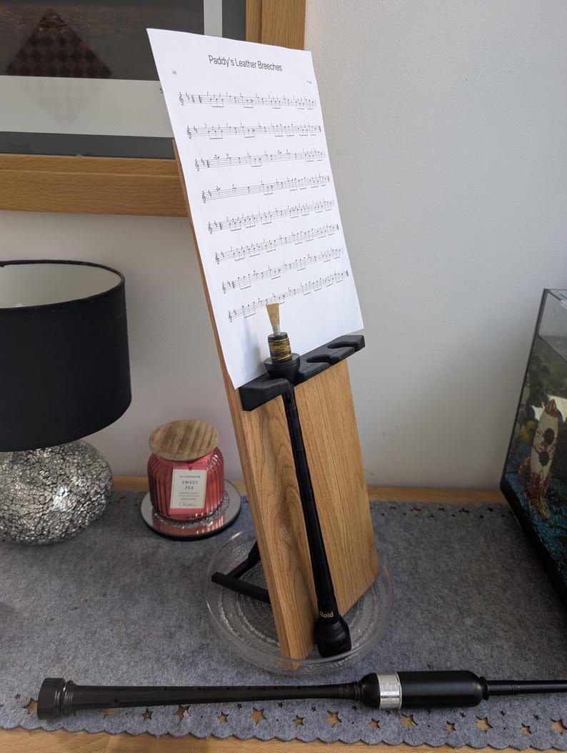May include: A black wooden stand with a sheet of music titled "Paddy's Leather Breeches" on top. A black wooden pipe is leaning against the stand. A black wooden pipe is lying on a gray surface.