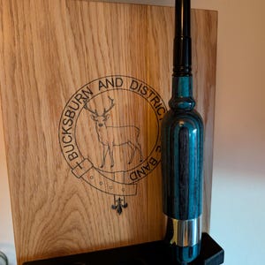 May include: A wooden wall mount with a black metal holder for a fishing rod. The wall mount has a logo that says "Bucksburn and District Band" with a deer and a fleur-de-lis design. The fishing rod is black with a blue and black patterned handle.