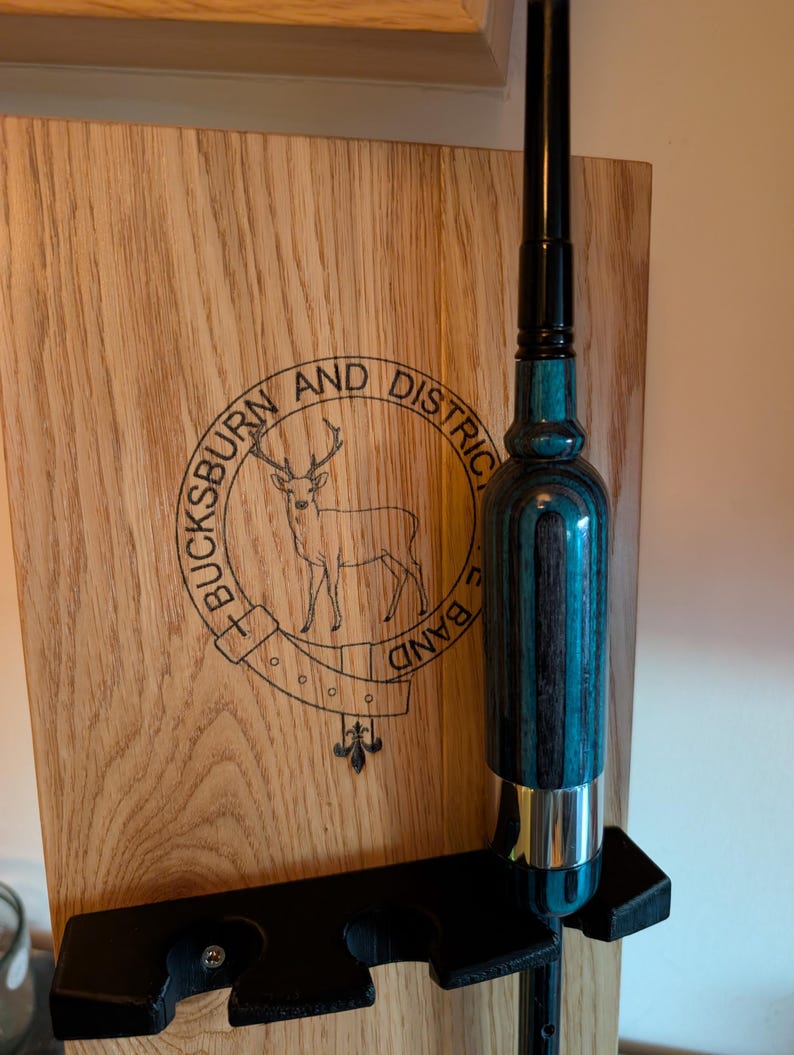May include: A wooden wall mount with a black metal holder for a fishing rod. The wall mount has a logo that says "Bucksburn and District Band" with a deer and a fleur-de-lis design. The fishing rod is black with a blue and black patterned handle.