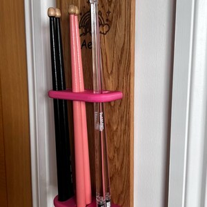 May include: A wooden wall-mounted rack with a pink plastic holder for drumsticks. The rack has a rainbow and cloud design with the text "Ae" engraved on it. There are three drumsticks in the holder, two pink and one black.