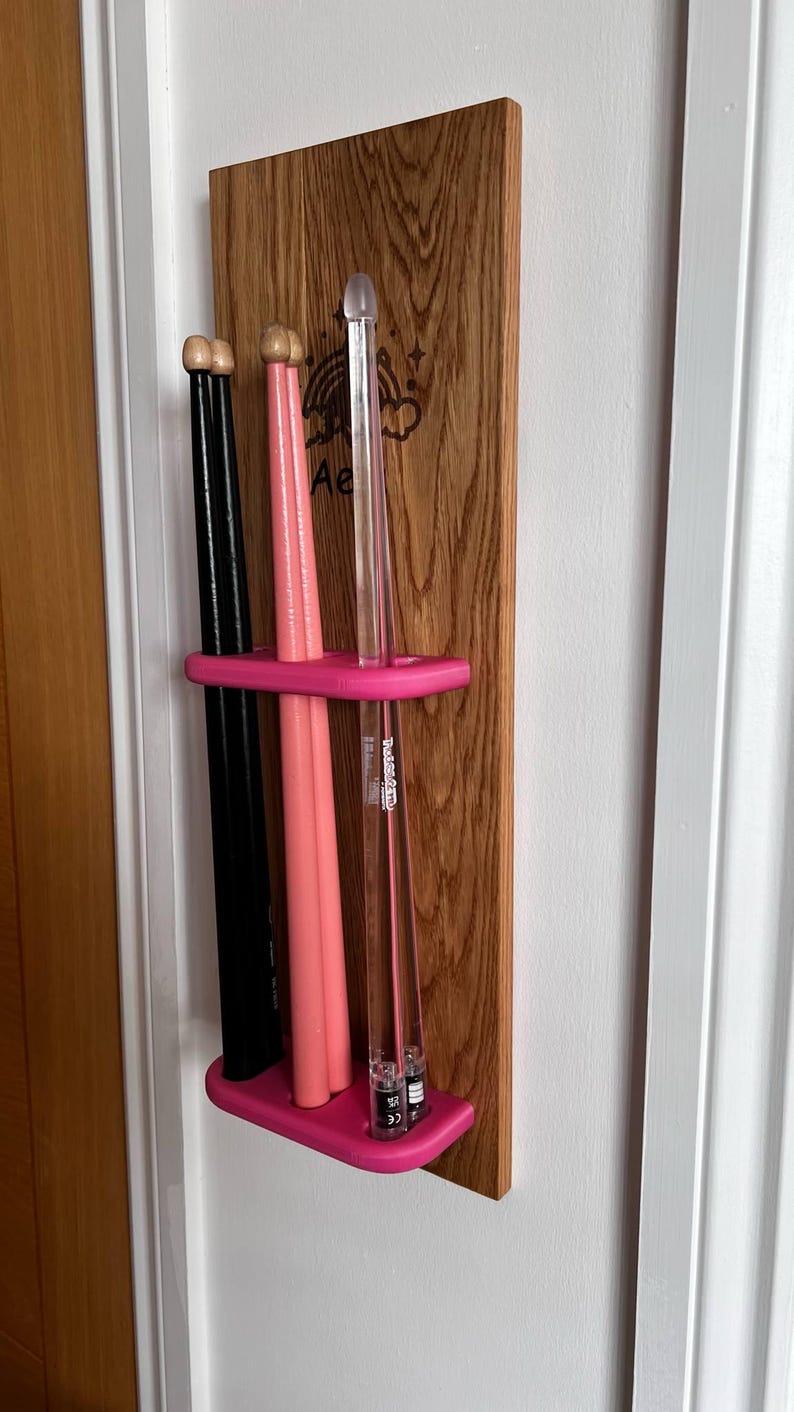 May include: A wooden wall-mounted rack with a pink plastic holder for drumsticks. The rack has a rainbow and cloud design with the text "Ae" engraved on it. There are three drumsticks in the holder, two pink and one black.