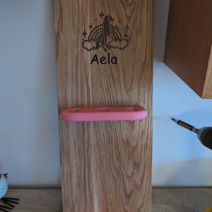 May include: A wooden wall-mounted organizer with two pink shelves. The top shelf has one slot and the bottom shelf has three slots. The wood is stained a light brown color. The organizer has a unicorn and rainbow design with the name "Aela" engraved on it.
