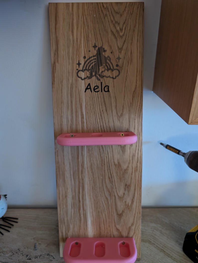 May include: A wooden wall-mounted organizer with two pink shelves. The top shelf has one slot and the bottom shelf has three slots. The wood is stained a light brown color. The organizer has a unicorn and rainbow design with the name "Aela" engraved on it.
