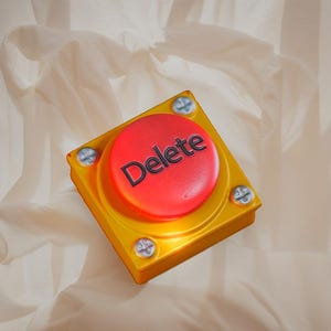 May include: A yellow square button with a red circular button in the center. The word "Delete" is printed in white on the red button. The button is secured with four silver screws. The background is a light beige fabric.