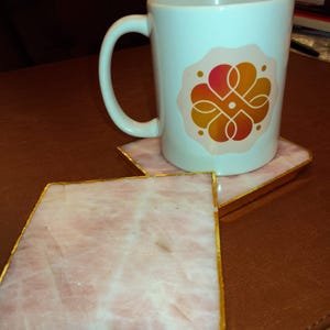 May include: A white ceramic mug featuring an orange and pink floral design, placed on a pink square coaster with gold edging. Another coaster is visible. The items are on a brown surface.