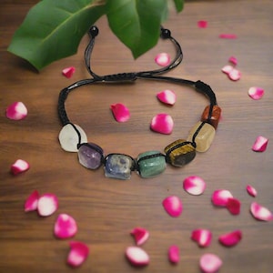 Puede incluir: Una pulsera de cordón negro con siete piedras preciosas pulidas de varios colores. Las piedras están dispuestas en fila e incluyen morado, verde y amarillo. La pulsera se exhibe sobre una superficie de madera con pétalos de rosa rosas esparcidos.