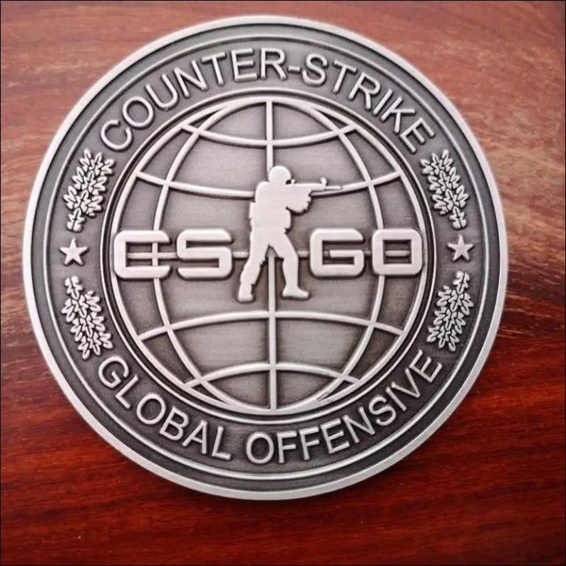 Może przedstawiać: Srebrna moneta z napisem "COUNTER-STRIKE GLOBAL OFFENSIVE" na obrzeżu. W centrum znajduje się globus z sylwetką żołnierza celującego z karabinu i literami "CS GO".