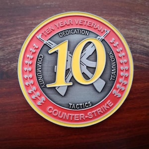 Może przedstawiać: Pamiątkowa moneta z czerwono-ż&oacute;łtą obw&oacute;dką, z napisem "TEN YEAR VETERAN" i "COUNTER-STRIKE". Liczba "10" jest wyraźnie wyświetlana na ż&oacute;łto, z skrzyżowanymi karabinami z tyłu. Moneta zawiera r&oacute;wnież słowa "DEDICATION", "COMMUNITY", "TEAMWORK" i "TACTICS".