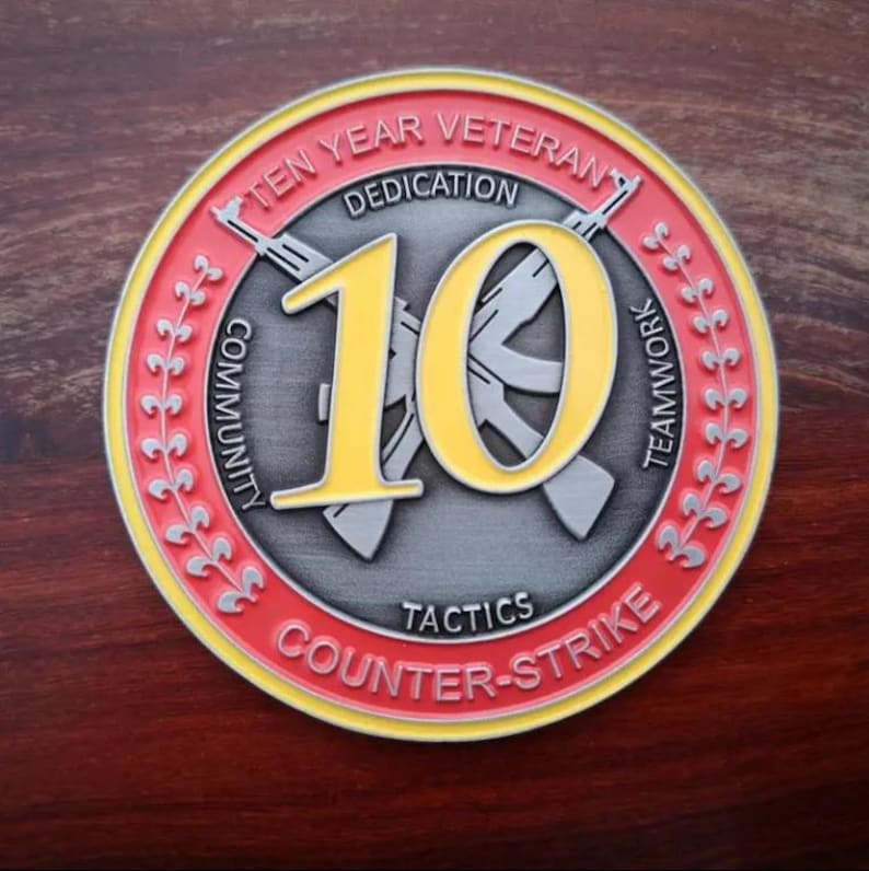 Może przedstawiać: Pamiątkowa moneta z czerwono-ż&oacute;łtą obw&oacute;dką, z napisem "TEN YEAR VETERAN" i "COUNTER-STRIKE". Liczba "10" jest wyraźnie wyświetlana na ż&oacute;łto, z skrzyżowanymi karabinami z tyłu. Moneta zawiera r&oacute;wnież słowa "DEDICATION", "COMMUNITY", "TEAMWORK" i "TACTICS".