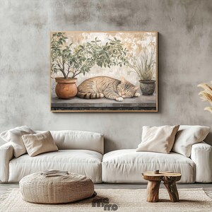 May include: A framed painting depicts a sleeping tabby cat next to potted plants. The artwork is set against a neutral background, with a light-colored sofa, woven ottoman, and wooden side table in the foreground. Dried ornamental grasses are in a vase.