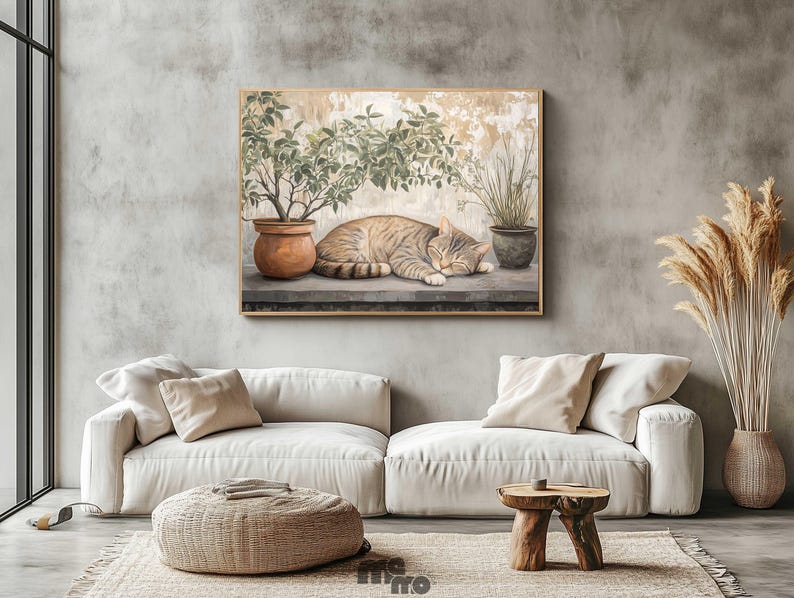 May include: A framed painting depicts a sleeping tabby cat next to potted plants. The artwork is set against a neutral background, with a light-colored sofa, woven ottoman, and wooden side table in the foreground. Dried ornamental grasses are in a vase.