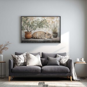 May include: A framed artwork depicts a sleeping tabby cat next to potted plants. The cat is tan and brown, and the plants are green. The artwork is displayed above a gray sofa with decorative pillows. A side table with a vase and a small table with books are also visible.