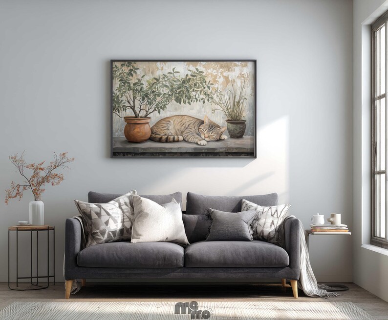 May include: A framed artwork depicts a sleeping tabby cat next to potted plants. The cat is tan and brown, and the plants are green. The artwork is displayed above a gray sofa with decorative pillows. A side table with a vase and a small table with books are also visible.